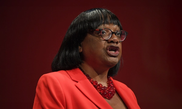 A woman with short black hair and glasses speaks passionately at a podium, wearing a bright red blazer and a matching necklace against a red backdrop.