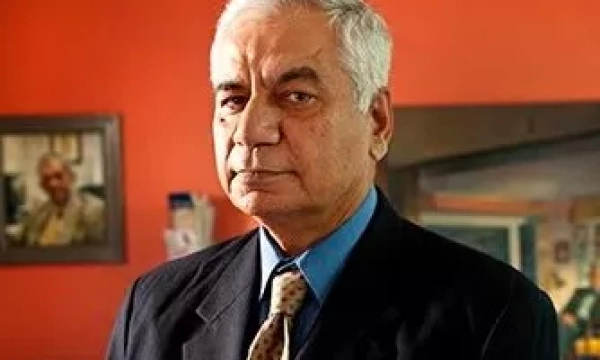 A man in a formal suit with a serious expression stands in front of an orange wall, with a photo of another person visible in the background.