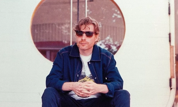 A man wearing sunglasses sits on wooden steps in front of a circular glass door, dressed in a casual outfit with a graphic t-shirt and jacket.