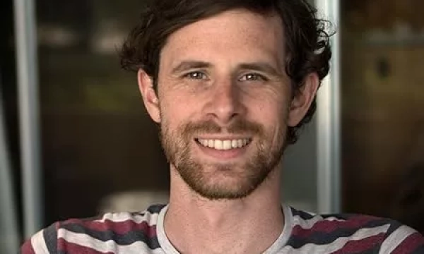 A smiling man with curly hair and a beard sits casually in a striped shirt, against a blurred background.