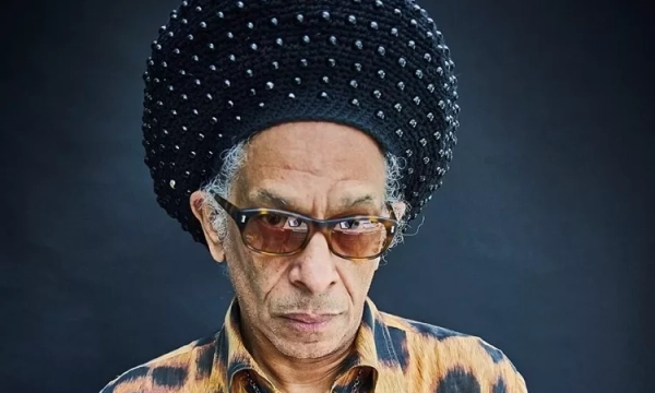 Portrait of a man with a large, beaded hairstyle and sunglasses, wearing a leopard-print shirt against a dark background.