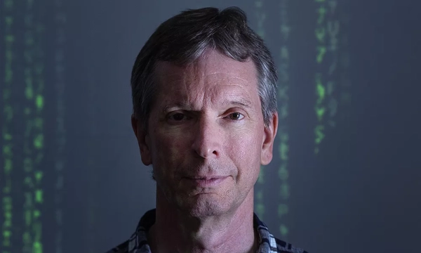 A middle-aged man with gray hair stands in front of a digital background featuring green code, evoking a tech-inspired atmosphere.