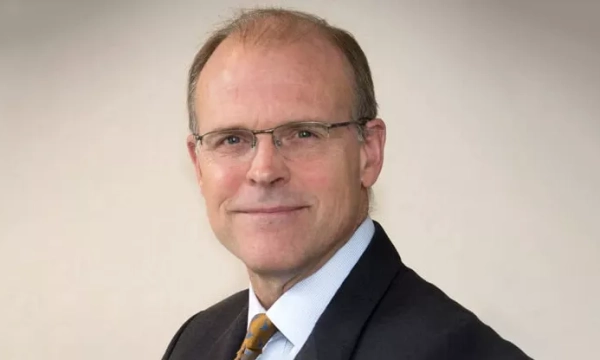A smiling man in a dark suit and light blue shirt, wearing glasses, poses confidently against a neutral background.