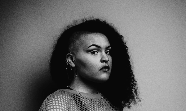 A portrait of a person with a half-shaved head, featuring curly hair on one side and a bold makeup look, captured in black and white.