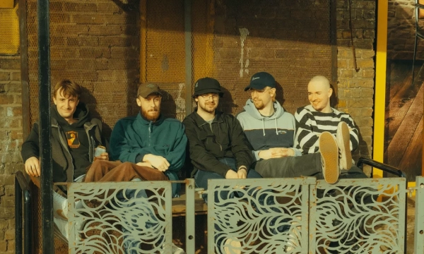 A group of five young men sitting together on a metal bench, smiling and relaxed, with a brick wall and industrial decor in the background.