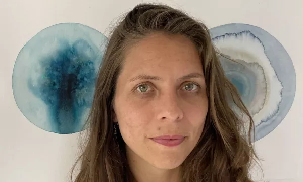 A woman with long, wavy hair stands in front of two circular blue and white watercolor artworks, displaying a serene expression.