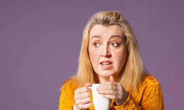 A woman with long blonde hair holds a white mug, looking anxious against a purple background.