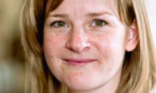 A smiling woman with light brown hair and green eyes, wearing a casual outfit, poses for the camera against a soft-focus background.