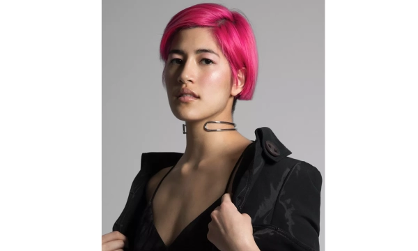 A person with pink hair wearing a black outfit poses confidently while holding their jacket, featuring a large safety pin accessory around their neck.