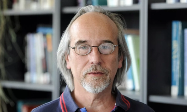 A middle-aged man with glasses and long, graying hair stands in front of a bookshelf filled with books.