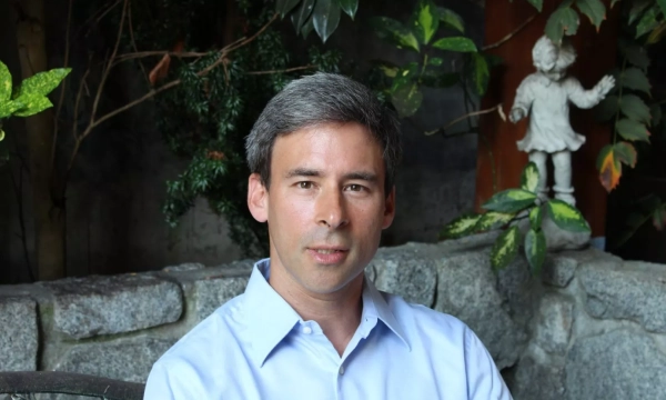 A man with gray hair sits on a stone bench, surrounded by greenery, with a decorative figure visible in the background.