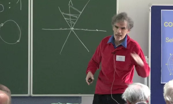 A presenter in a red sweater gestures while explaining a concept in front of a chalkboard with geometric drawings.