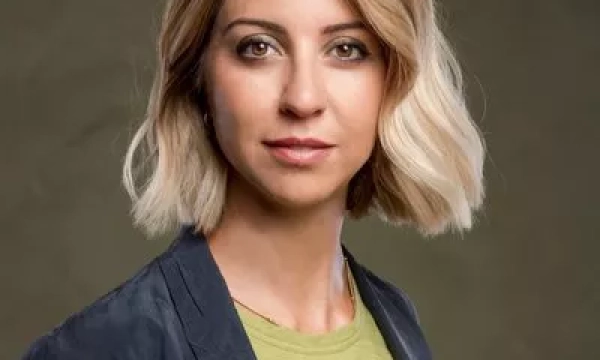 A woman with shoulder-length blonde hair, wearing a green shirt and navy blazer, poses against a neutral background, looking directly at the camera.