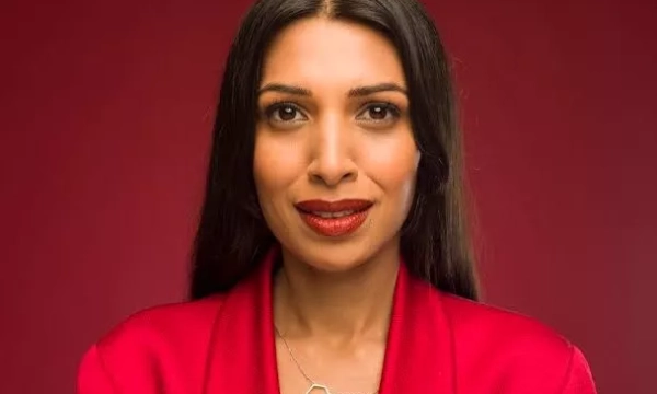 A woman with long, straight black hair and red lipstick poses confidently in a red blazer against a solid red background.