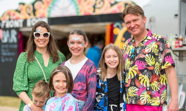 A cheerful family poses at a vibrant outdoor event, featuring colorful clothing and face paint, with a festive backdrop and a bar sign in the background.