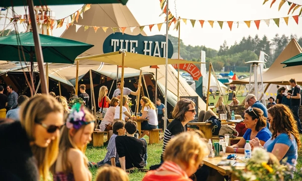 People enjoying a lively outdoor festival with colorful tents, decorative bunting, and picnic tables. A diverse crowd engages in conversation and activities.
