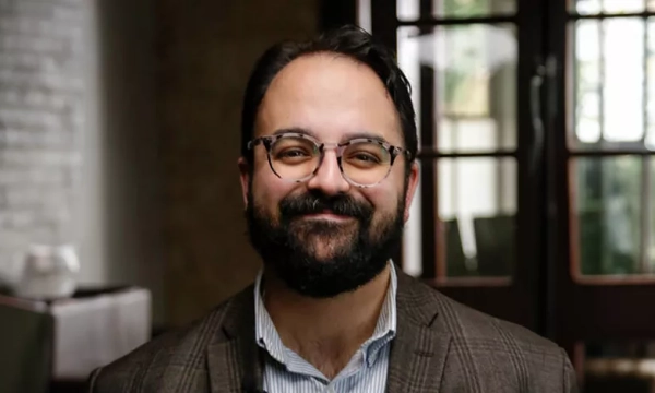 A smiling man with glasses and a beard, wearing a blazer, poses in a well-lit interior setting with large windows in the background.