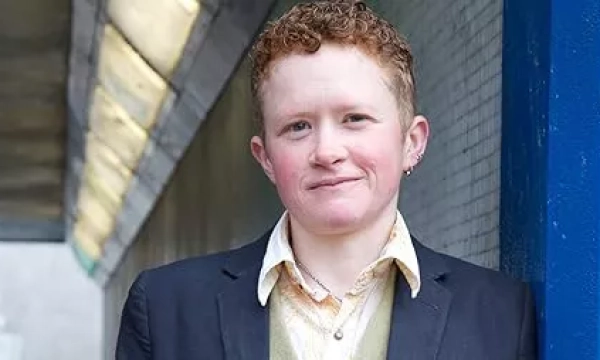 A person with short, curly hair wearing a blazer and an open-collared shirt stands in a modern urban setting, against a backdrop of a tiled wall and overhead lights.