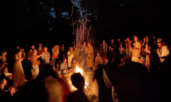 A vibrant gathering around a bonfire at night, with people dancing and celebrating, surrounded by trees and a warm glow from the flames.