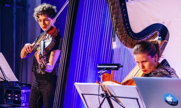 Two musicians perform together, one playing the violin and the other on a harp, with colorful stage lighting.