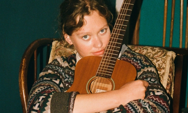 A young person with wavy hair sits on a chair, hugging an acoustic guitar, dressed in a patterned oversized sweater, exuding a contemplative mood.
