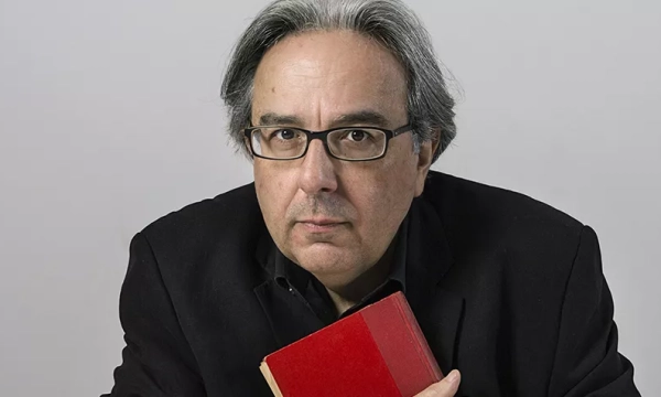 A middle-aged man with glasses, dressed in a black jacket, holding a red book and looking directly at the camera against a plain background.