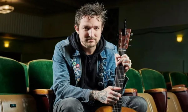 A man with tattoos and a denim jacket sits in a theater, holding an acoustic guitar, surrounded by empty green seats.