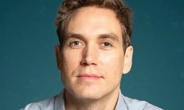 A close-up portrait of a man with light brown hair, wearing a button-up shirt against a teal background, displaying a neutral expression.