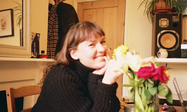 A smiling woman in a black sweater sits at a table decorated with a vase of colorful flowers, surrounded by a cozy indoor setting.