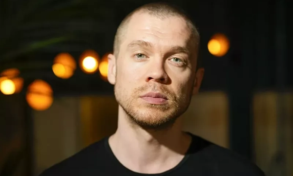 A close-up portrait of a man with a shaved head and intense expression, set against a backdrop of blurred warm lights.