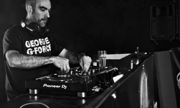 A DJ focused on the turntable, wearing a "George G-Force" t-shirt, with club equipment and a glass nearby, illuminated in a dim setting.