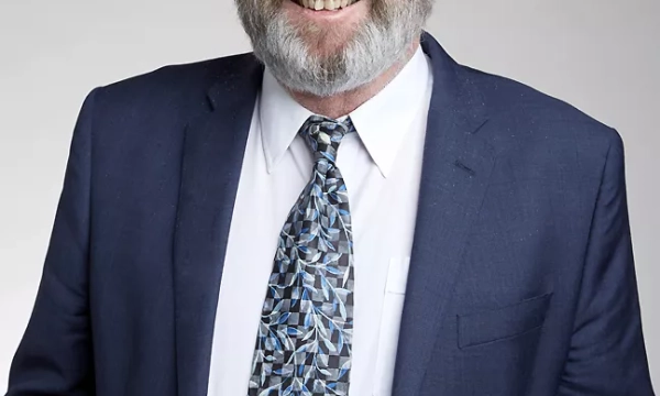 A man with gray hair and a beard wearing glasses, a dark suit, a white shirt, and a patterned tie, smiling in front of a simple background.