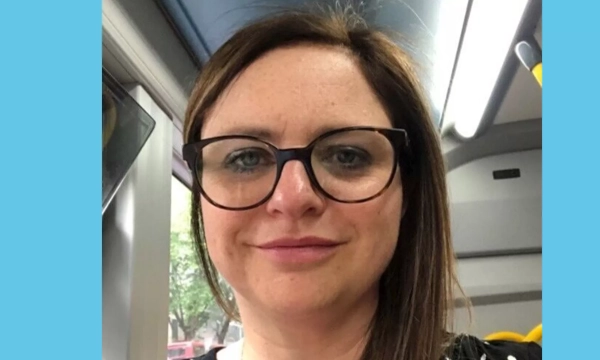 A woman with long brown hair and glasses smiles for a selfie on a bus, showcasing a casual environment.