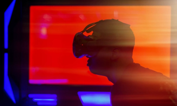 A silhouetted person wearing a virtual reality headset stands against a vibrant, illuminated background with red and orange hues.