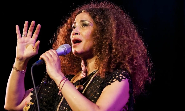 A woman with curly hair passionately sings into a microphone, wearing a black dress and jewelry, against a dark background with stage lighting.