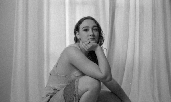 A person squatting near a window with sheer curtains, wearing a layered outfit and black boots, with a thoughtful expression. The image is in black and white.