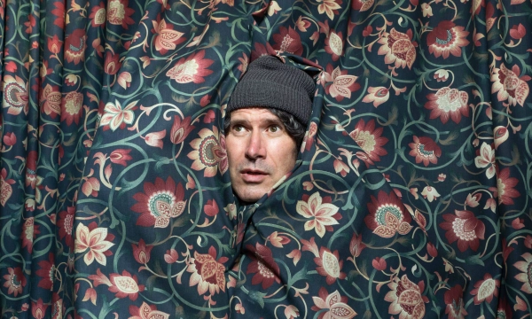 A man partially wrapped in a floral-patterned curtain, wearing a beanie, with a surprised expression on his face.