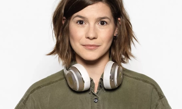 A young woman with shoulder-length brown hair and wearing a green shirt, posing with headphones around her neck, against a plain white background.