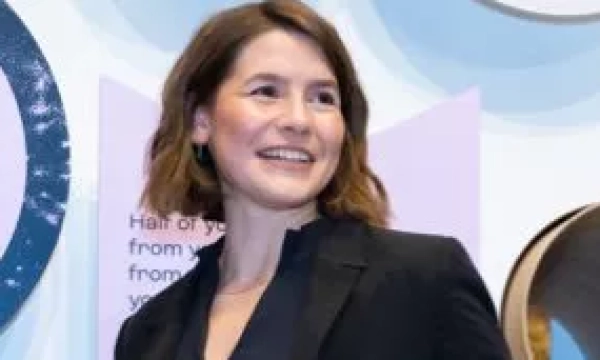 A woman with shoulder-length brown hair, wearing a black blazer, smiles while standing in front of informational displays at an event.