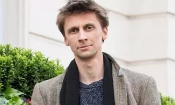 A man standing outside near greenery, wearing a casual outfit with a scarf, looking directly at the camera.