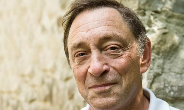 A smiling older man with light brown hair stands against a textured stone background, conveying warmth and approachability.