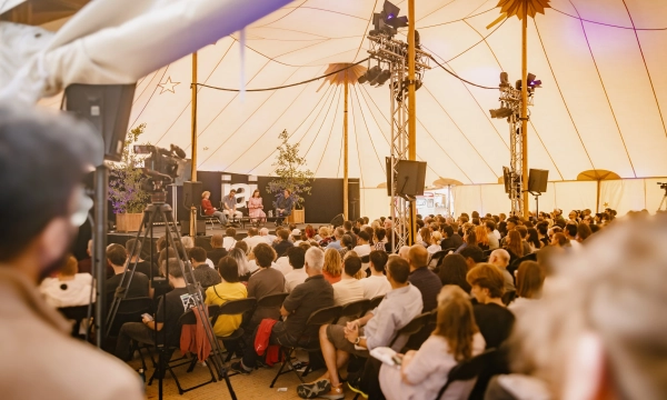 A large audience seated under a tent, watching speakers on stage, with lights and cameras capturing the event.