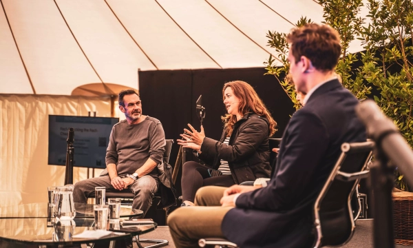 A panel discussion featuring three individuals seated in modern chairs, engaged in conversation under a tent with plants and a screen in the background.
