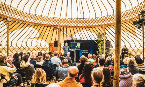 A speaker presents to an audience inside a large yurt, with a screen displaying visuals in the background. The audience appears engaged and attentive.
