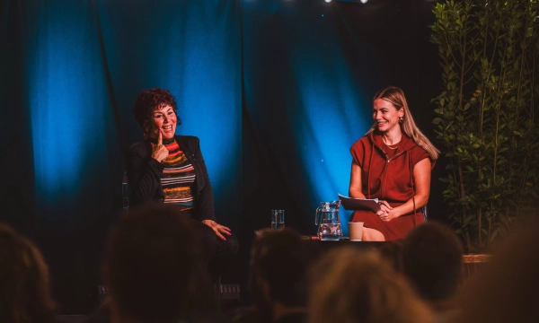 A woman with curly hair and colorful attire smiles while sitting beside another woman in a red dress, both engaged in a discussion on stage.