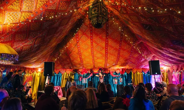 A vibrant, decorated tent filled with people watching performers in colorful attire, illuminated by string lights and a hanging lantern.