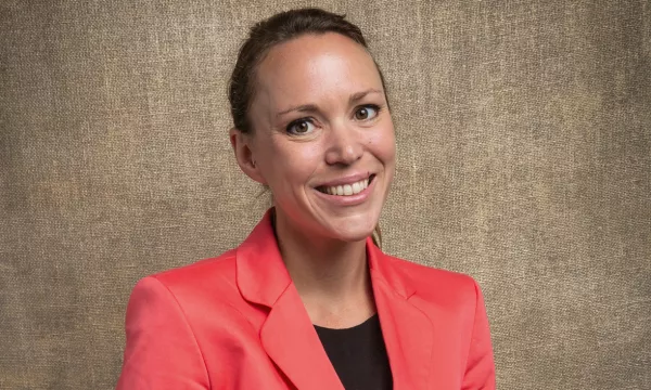 A smiling woman wearing a coral blazer poses against a textured beige background. Her expression is warm and approachable.