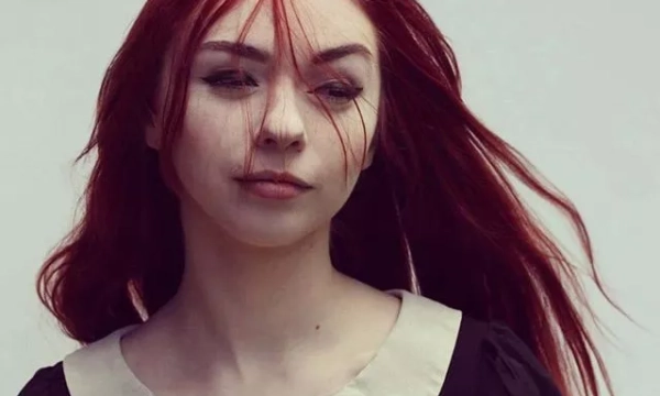 A young woman with long red hair and a vintage-style black dress featuring a white collar, gazing thoughtfully with wind-blown strands of hair.