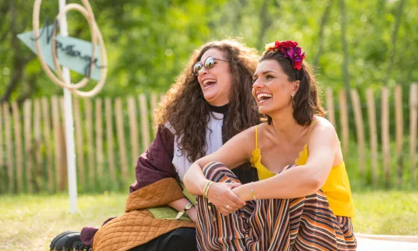 Two women laughing together outdoors, dressed in vibrant, colorful outfits, enjoying a sunny day in a grassy area with a wooden fence in the background.