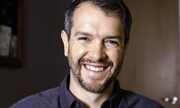 A smiling man with short, dark hair and a beard, wearing a dark shirt, seated in a well-lit interior setting.